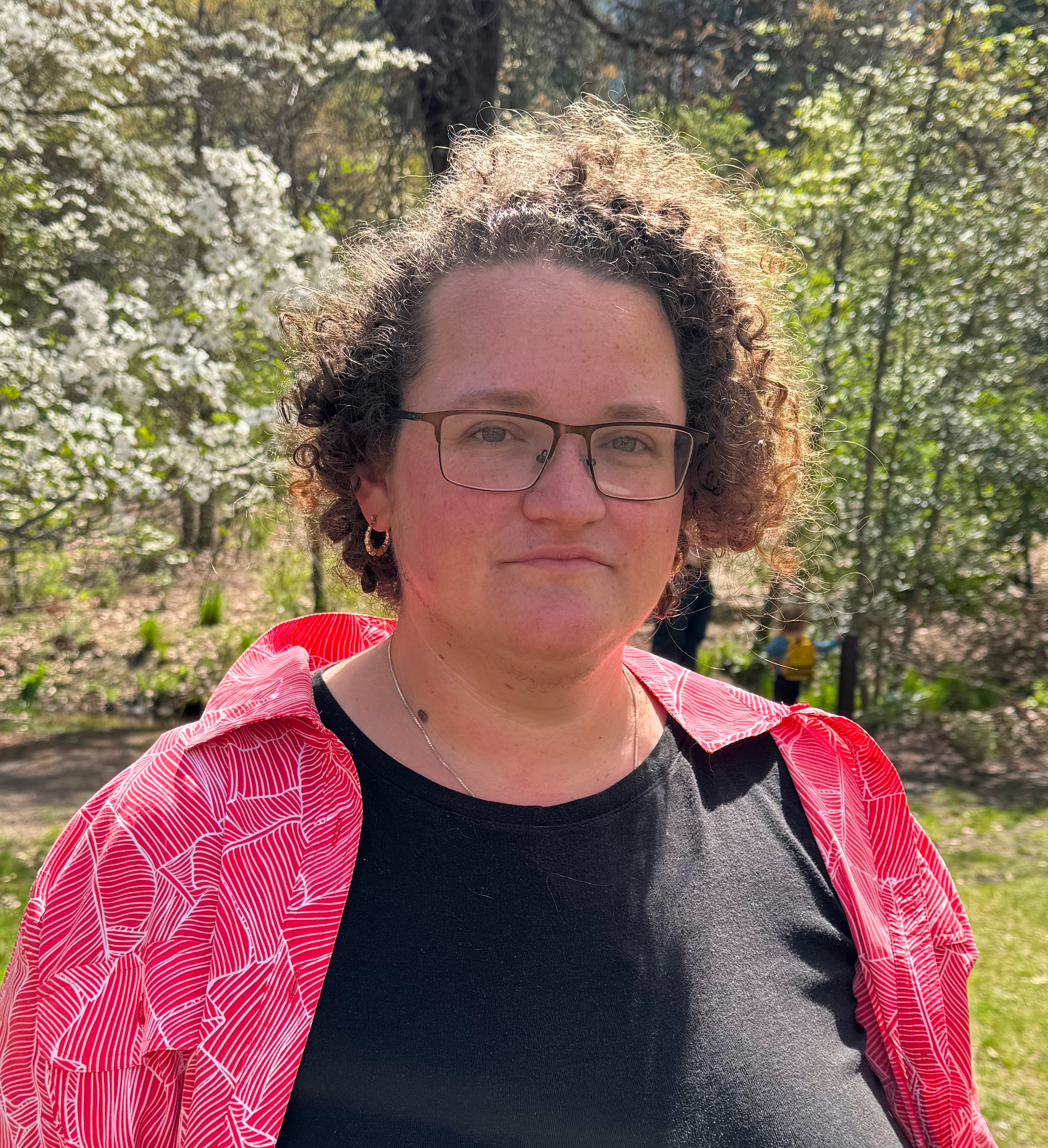 Headshot of Caroline Taymor, a white curly haired person in a black shirt with red overshirt in front of dogwoods